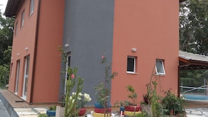 A two-story building with a red and grey facade, a small window, and a paved entrance area with potted plants. - Mini Club with Biribol (a Brazilian beach volleyball game) (Bertioga)
