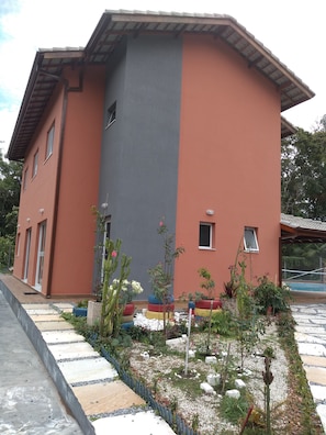 A two-story building with a red and grey facade, a small window, and a paved entrance area with potted plants. - Mini Club with Biribol (a Brazilian beach volleyball game) (Bertioga)