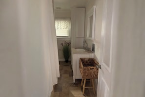 Un pasillo estrecho con una puerta blanca, una pequeña cesta de mimbre sobre un taburete y una planta en maceta.