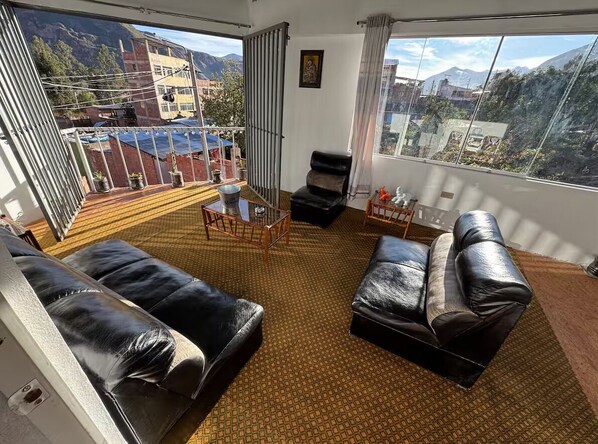 A room with a large window, a black leather sofa, a small table, and a view of mountains.