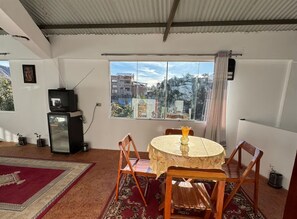 Ein Zimmer mit einem runden Tisch, vier Stühlen, einem Fernseher und einem Fenster mit Blick auf Gebäude.