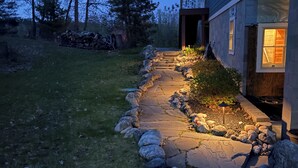 A stone pathway leading to a house with a porch, surrounded by grass and rocks.