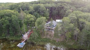 A house surrounded by dense forest, a dock with a boat, and a small bridge.