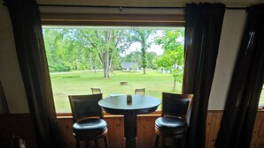 A room with a round table and four chairs, a window with curtains, and a view of a grassy area with trees. - Snowmobile Sanctuary in the Charming Village of Crivitz (Crivitz)