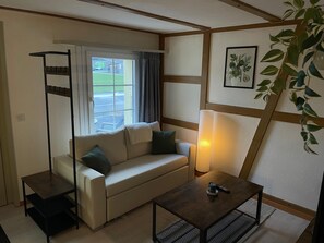 A cozy room with a sofa, a small table, a lamp, a window with curtains, a plant, and a framed picture on the wall.