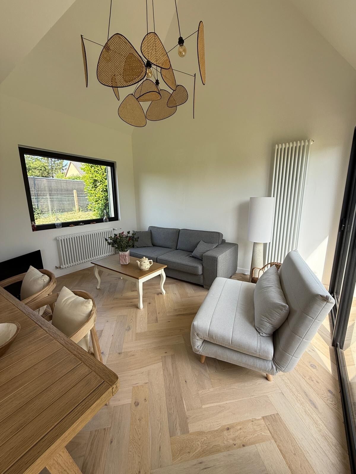 A modern living room with a grey sofa, a white armchair, a wooden dining table, and a hanging light fixture.