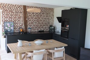 A modern kitchen with a brick wall, black appliances, a wooden dining table, and white chairs. - Family home in the countryside, 25 km from Rouen (Auzouville-sur-Ry)