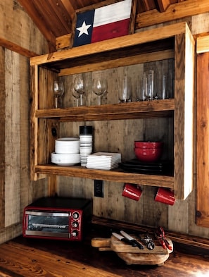 A wooden kitchen with a Texas flag, a red toaster oven, stacked white plates, red mugs, and a wooden cutting board with knives.