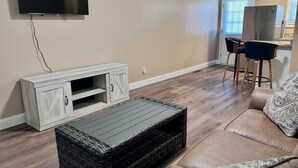 A living room with a flat-screen TV mounted on the wall, a wooden entertainment unit, a wicker coffee table, a leather sofa, and a kitchen area with a refrigerator and bar stools. - In-Town Gem • Huge Yard • Near Golf, Food & attractions (Crossville)