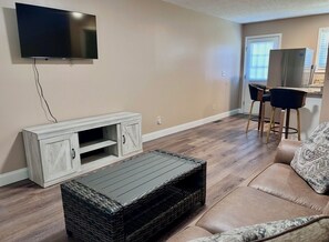 A living room with a flat-screen TV mounted on the wall, a wooden entertainment unit, a wicker coffee table, a leather sofa, and a kitchen area with a refrigerator and bar stools.