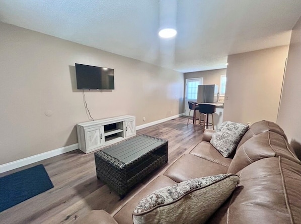Un salon avec un canapé en cuir marron, un téléviseur à écran plat fixé au mur, un meuble de rangement blanc et un coin repas comprenant une table et des chaises.