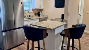 A compact kitchen with a refrigerator, two bar stools, a countertop, and a window with blinds. - In-Town Gem • Huge Yard • Near Golf, Food & attractions (Crossville)