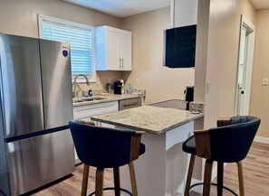 A compact kitchen with a refrigerator, two bar stools, a countertop, and a window with blinds.