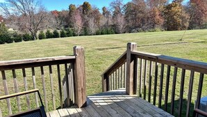 A wooden deck with a metal bench and a railing, overlooking a grassy field with trees. - In-Town Gem • Huge Yard • Near Golf, Food & attractions (Crossville)