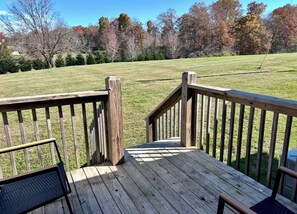 A wooden deck with a metal bench and a railing, overlooking a grassy field with trees.