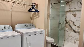 Uma área de lavanderia com duas máquinas de lavar e secadoras brancas, um box de chuveiro envidraçado e parede de chuveiro revestida em mármore.