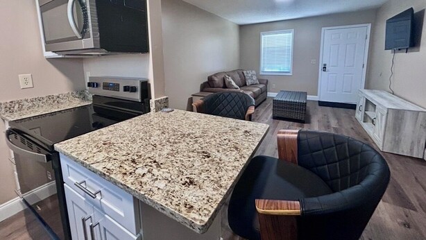A compact kitchen with a microwave, stove, and dishwasher. There is a dining area with a granite countertop and a black chair. A living area with a couch and a television is visible. - In-Town Gem • Huge Yard • Near Golf, Food & attractions (Crossville)