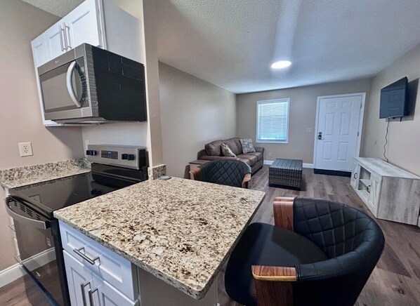 A compact kitchen with a microwave, stove, and dishwasher. There is a dining area with a granite countertop and a black chair. A living area with a couch and a television is visible.