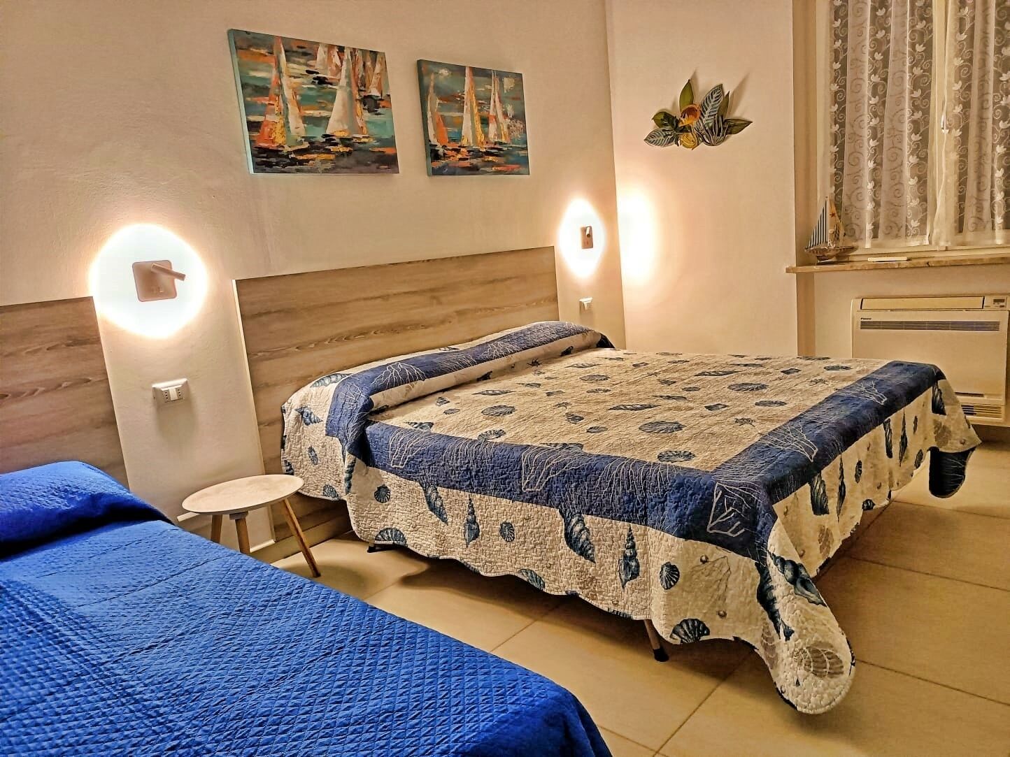 Una habitación de hotel con cama, dos mesitas de noche, luces fijadas en la pared, una mesa redonda pequeña y dos cuadros.