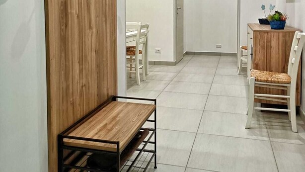Un interior moderno con una pared de madera, un banco de metal, una zona de comedor con sillas blancas y un televisor fijado a la pared.