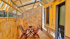 Un patio techado al aire libre con una mesa y sillas, piso de baldosas y una puerta con ventanal.