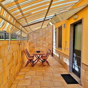 Eine überdachte Terrasse mit Tisch und Stühlen, ein Fliesenboden und eine Tür mit Fenster.