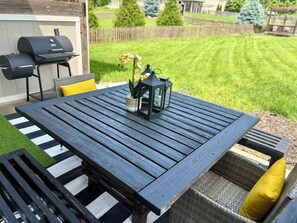 Ein Holztisch auf einer Terrasse, mit einer Laterne und Blumen, umgeben von Korbmöbeln und einem Streifen Teppich.