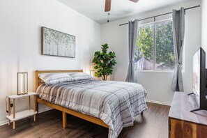 Une chambre à coucher avec un lit, une table de chevet, une plante, une fenêtre avec des rideaux et un tableau au mur.