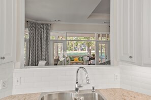 A kitchen with a window view of a patio area, a stainless steel sink, and white tiled walls. - Beautiful Renovated 3BR/3BA Retreat Free Golf, Riverfront Pools & Resort Living (Okatie)