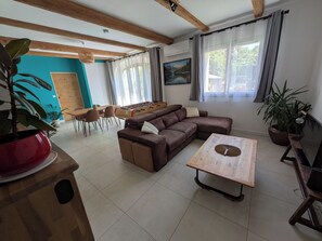 Un salon comprenant un canapé d’angle, une table basse en bois, un coin repas avec des chaises, une plante en pot rouge et une grande fenêtre avec des rideaux.