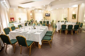 A banquet hall with multiple round tables, green upholstered chairs, white tablecloths, and a television mounted on the wall.