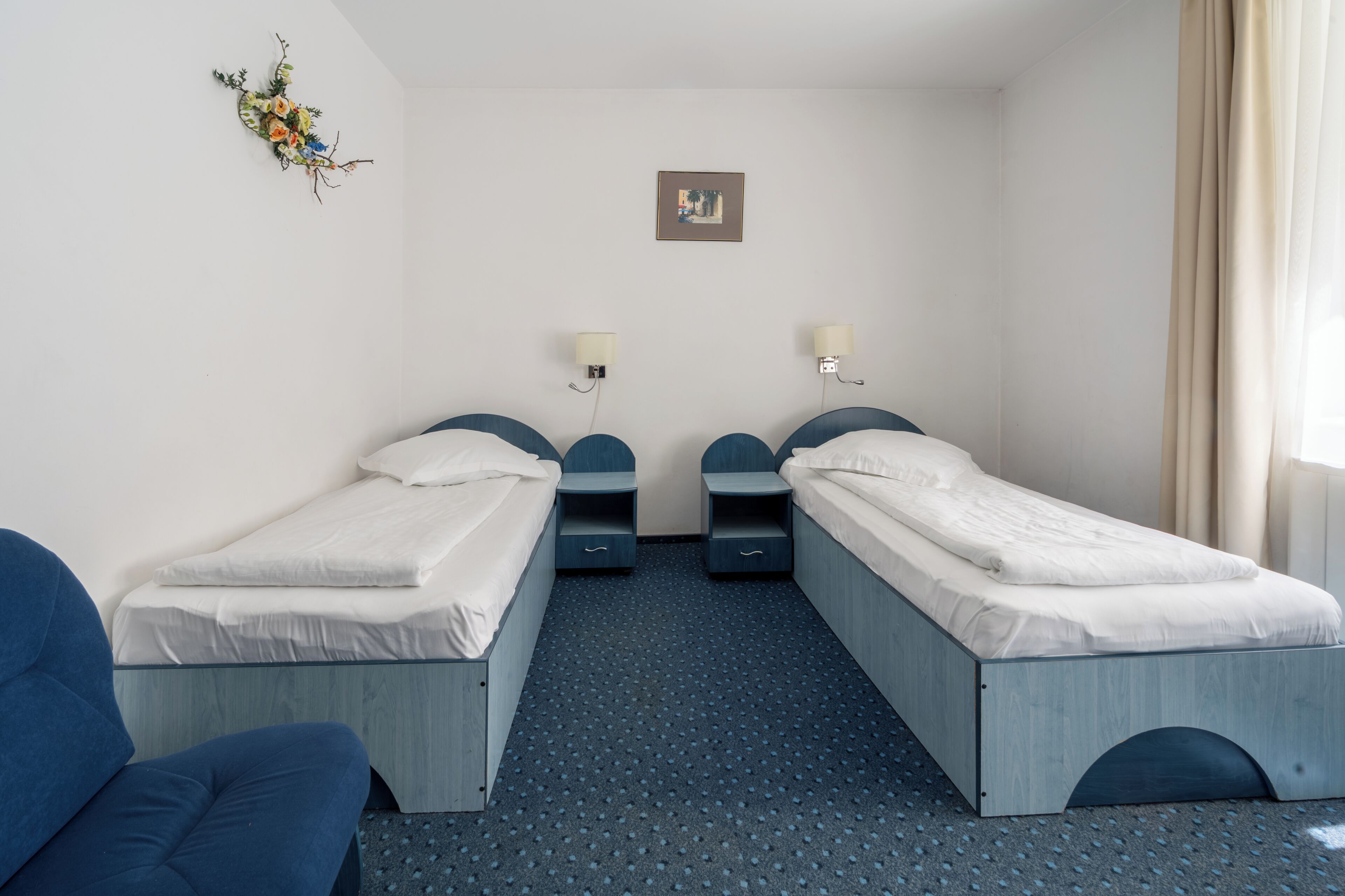 A hotel room with two single beds, a small desk, a chair, a wall-mounted lamp, and a framed picture.
