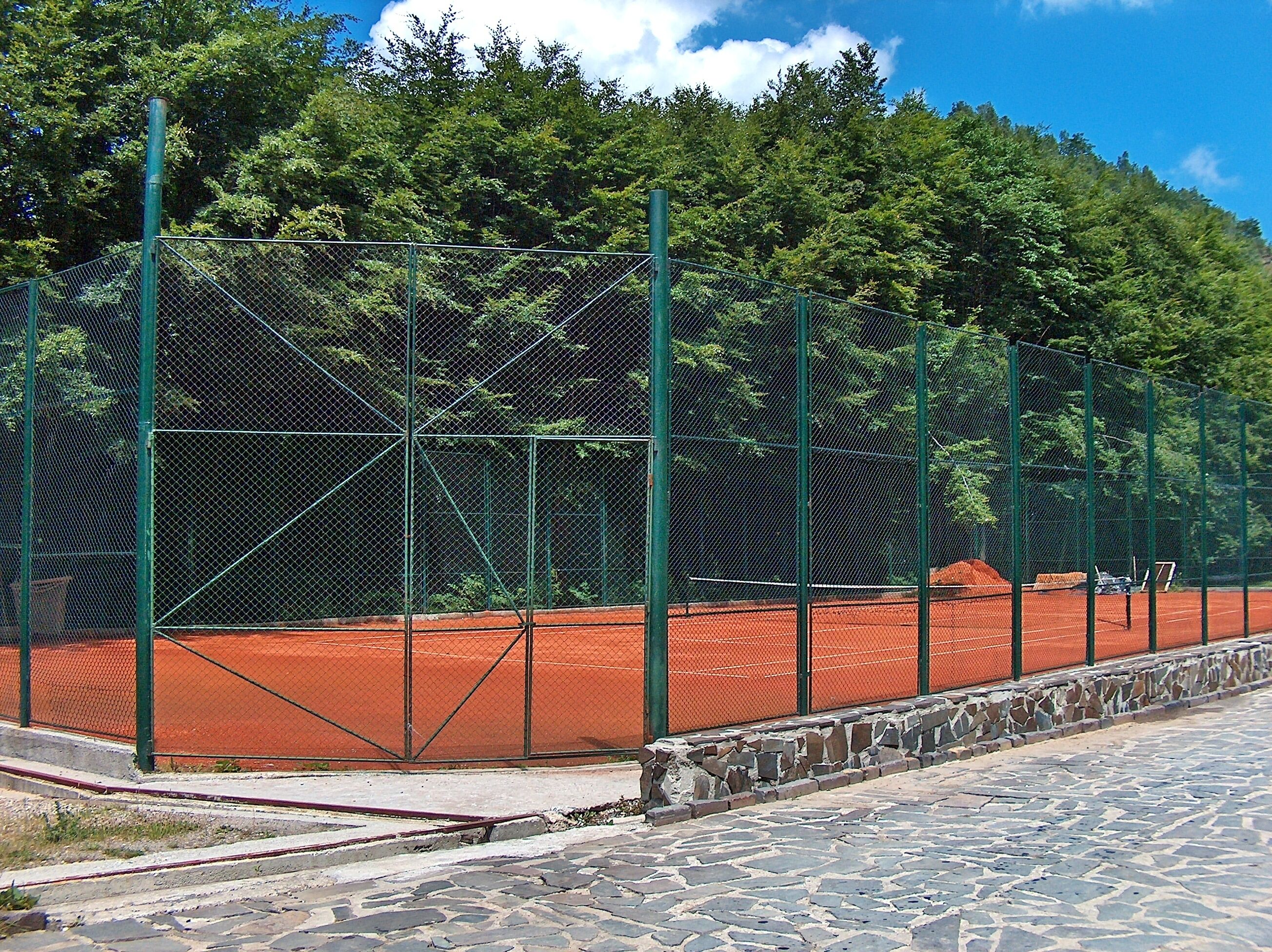 A tennis court with a green fence, surrounded by trees and a stone wall.
