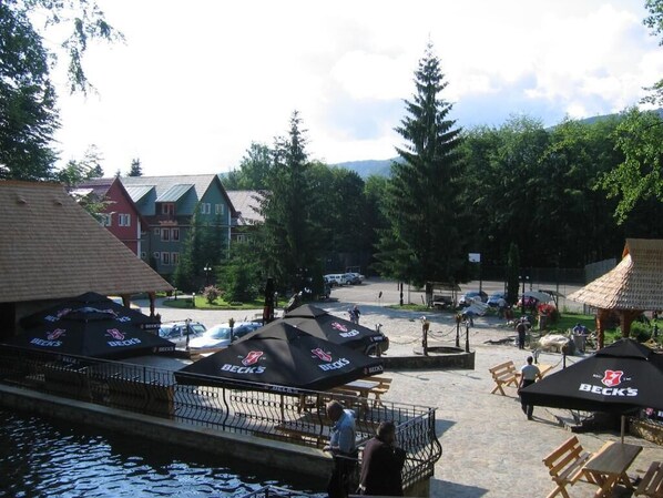 A lakeside outdoor seating area with umbrellas, wooden benches, and a view of buildings and trees.