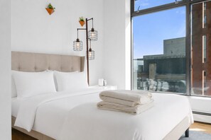 Ein modernes Schlafzimmer mit einem großen Bett, einem Nachttisch, einer Lampe und Blick auf die Stadt durch ein großes Fenster.