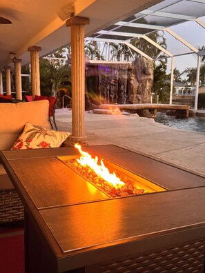 A fire feature with a stone surround, a seating area with a cushion, and a water feature in the background. - Waterpark Style Pool With Hot Tub in Grotto With Waterfall and Bridge Over Pool (Cape Coral)
