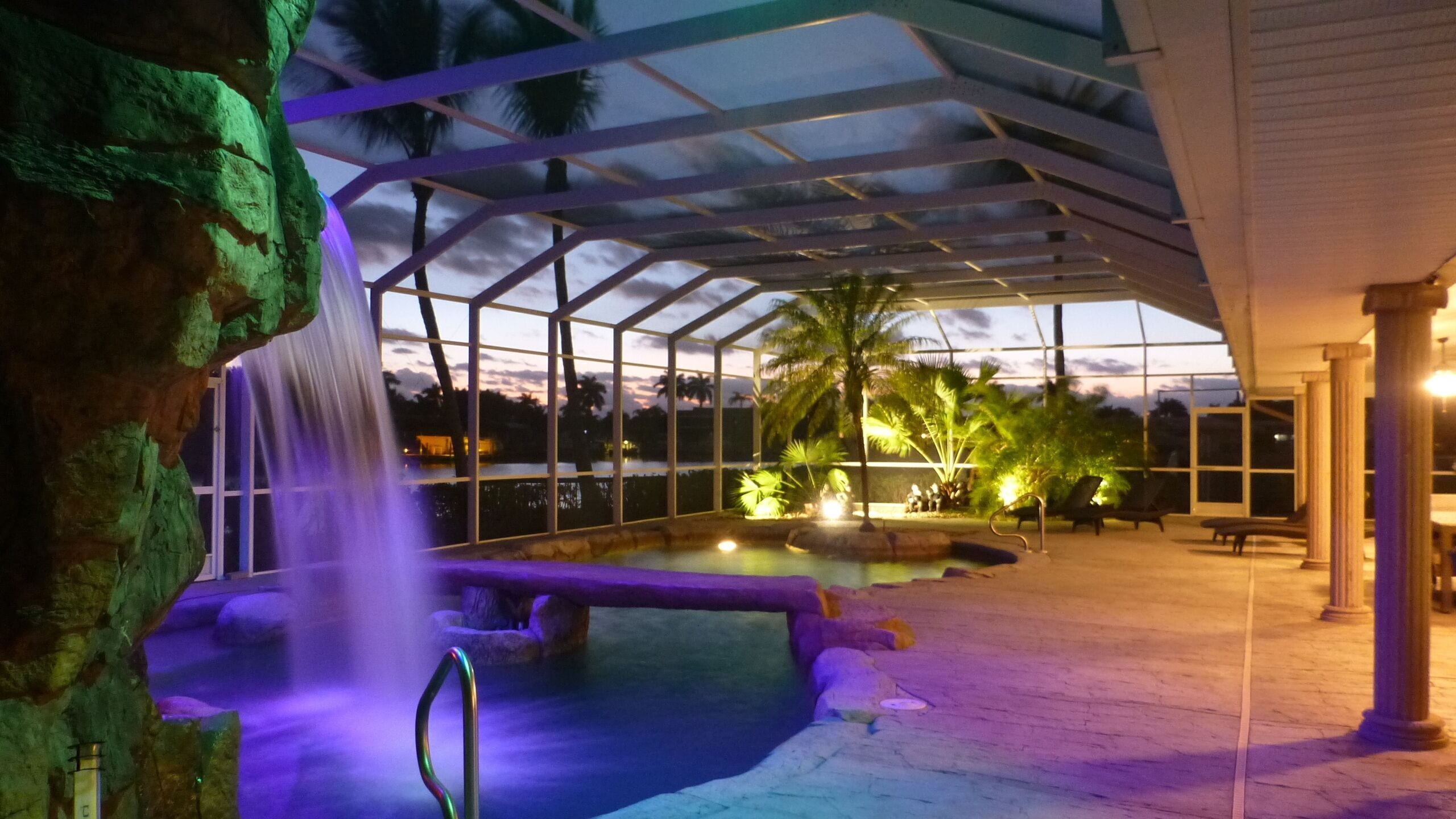 An outdoor pool area with a waterfall feature, surrounded by palm trees and a covered seating area.