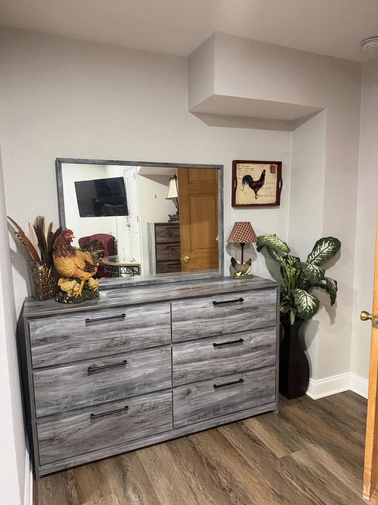 Een hoekkamer met een grijze houten dressoir, een grote spiegel, een afbeelding van een haan in een lijst, een lamp en een plant in pot.