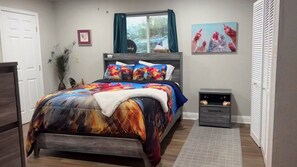 A bedroom with a bed, a bedside table, a window with curtains, a wall-mounted artwork, and a door.
