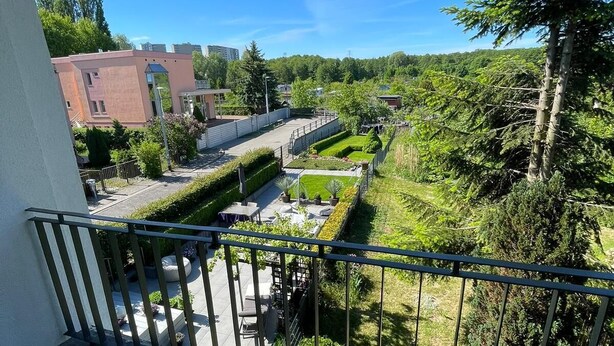 Un balcón con vistas a un jardín ajardinado, un edificio al fondo y un cielo azul despejado.