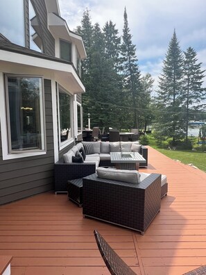 A modern outdoor deck with a sectional sofa, a glass-top table, and a view of a lake and forest.