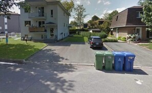 Rua residencial com casas, um carro estacionado e três latas de lixo.
