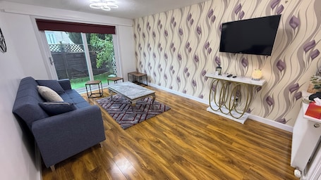 A living room with a sofa, a coffee table, a TV mounted on the wall, and a patterned wallpaper.. Clean & Spacious 2BR Home Near Glasgow Airport and Loch Lomond