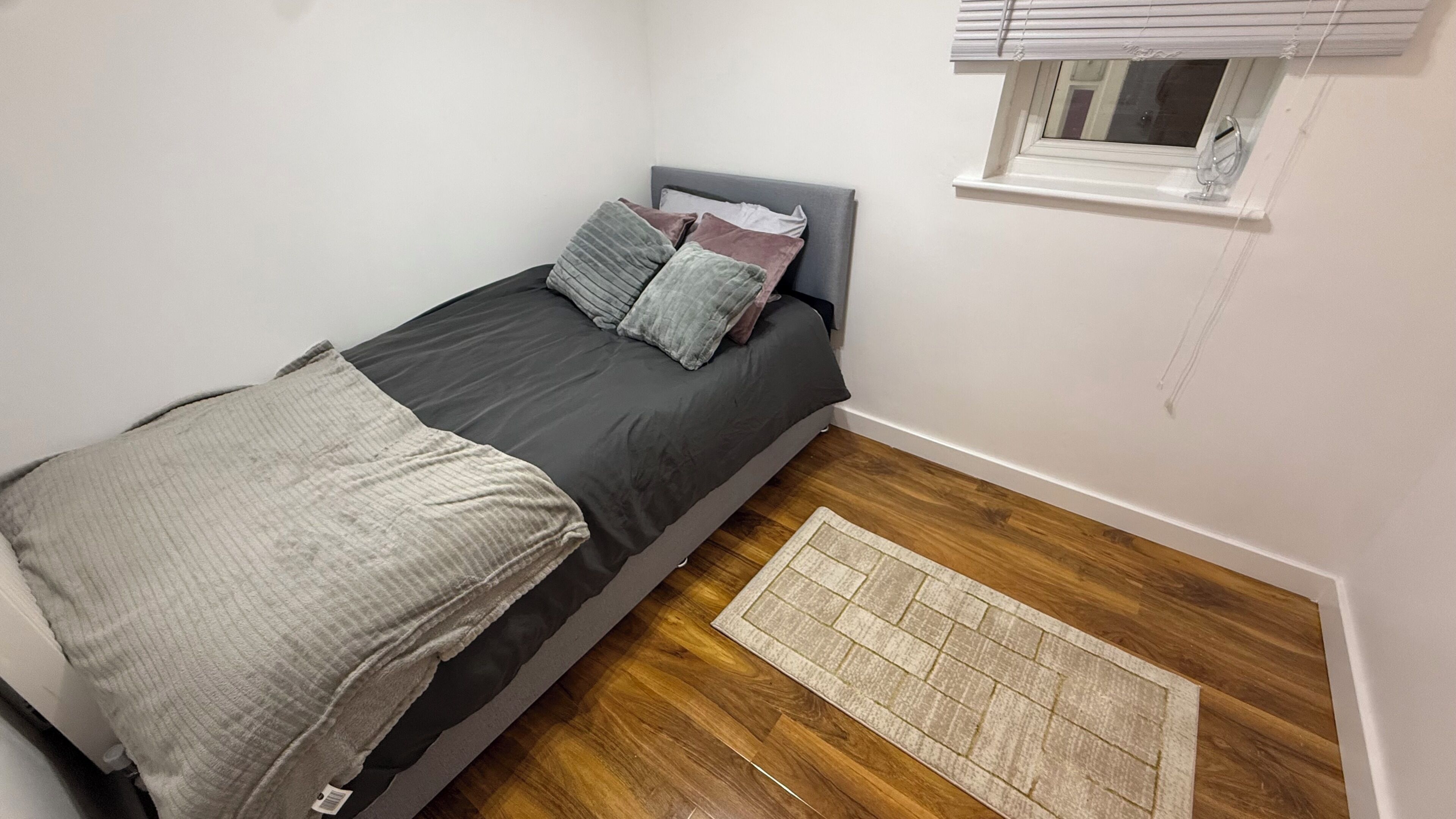 A small, single-bed room with a grey headboard, a window with blinds, a wooden floor, and a small rug.