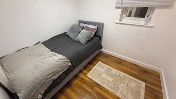 A small, single-bed room with a grey headboard, a window with blinds, a wooden floor, and a small rug.