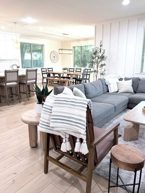 Un salon moderne avec un canapé gris, des chaises en bois, une table à manger et une grande fenêtre.
