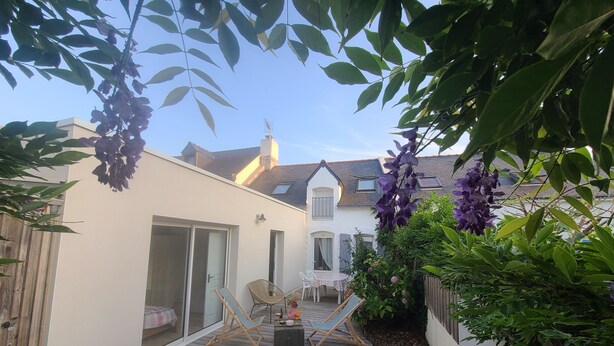 A wooden deck with two chairs, a table, and a sliding glass door leading to a white building with a sloped roof. - Charming town house, 10min walk to the beach (saint gildas de rhuys)