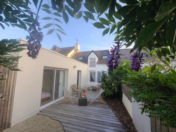 A wooden deck with two chairs, a table, and a sliding glass door leading to a white building with a sloped roof. - Charming town house, 10min walk to the beach (saint gildas de rhuys)