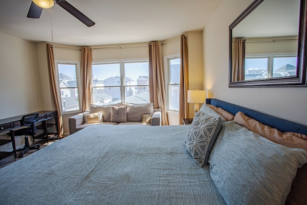 Une chambre d’hôtel avec un grand lit, un bureau, une chaise, un ventilateur de plafond et une vue sur les immeubles par les fenêtres.