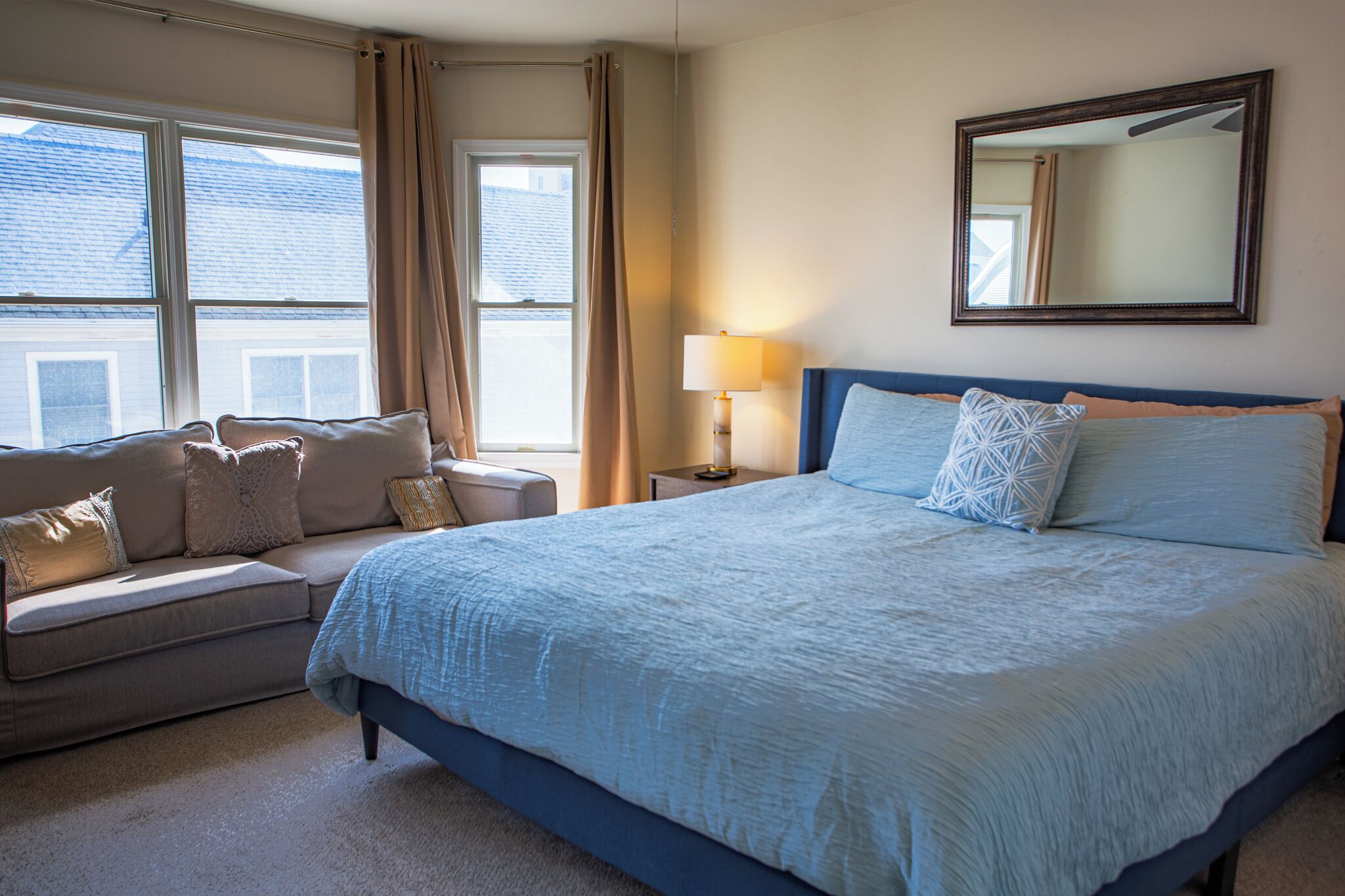 A bedroom with a bed, a sofa, a lamp, a mirror, and windows.
